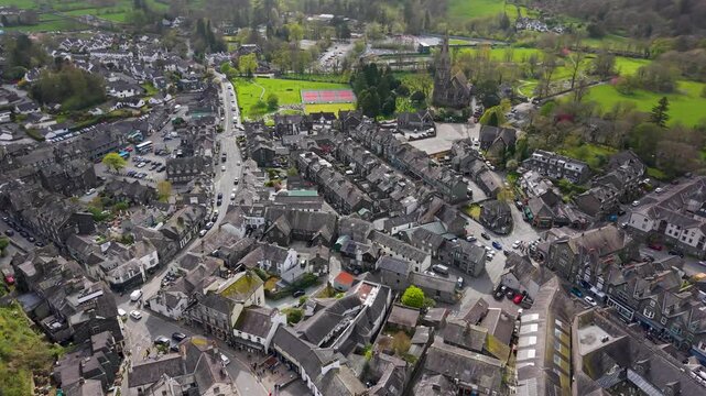 A sweeping left orbital aerial shot over Ambleside town centre, revealing stone buildings, winding streets, St Mary's Church, and lush green fields beyond. Lake District, Cumbria.