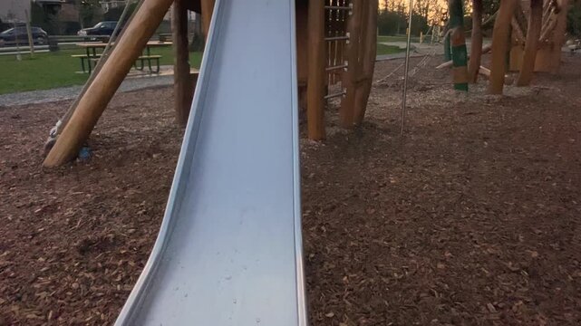 Metal playground slide pathway stretching downward towards wood chip ground cover captured by moving camera POV sliding under cloudy twilight sky near suburban neighborhood houses