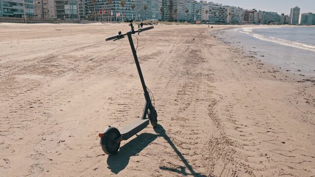 An electric scooter stands on a sandy beach with city buildings and the ocean in the background, perfect for travel and urban mobility concepts.