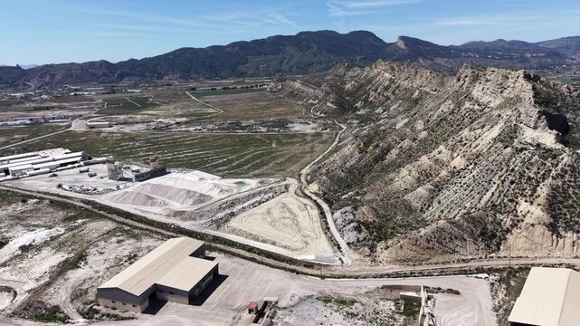 A Tracking and turning drone shot showcasing a large cement factory and gravel business with massive piles of raw materials and industrial warehouses.