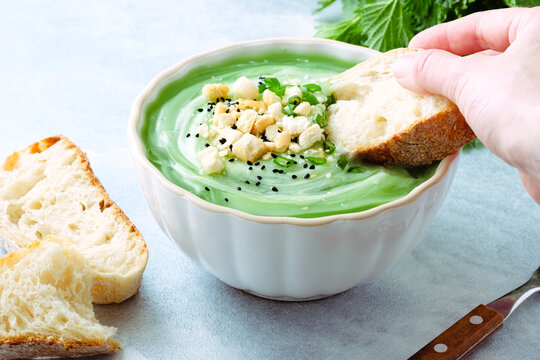 A hand dips a piece of bread into a bowl of green soup topped with herbs and crackers during lunchtime on a simple table. Fresh herbs are nearby. Healthy food.