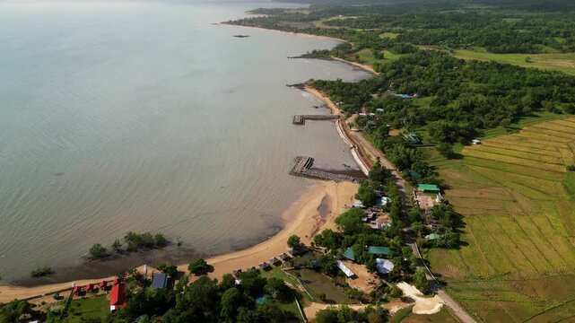 Aerial flyover of a coastal barangay village with sandy shoreline, farmland, and calm sea in San Lorenzo, Guimaras Island, Philippines during daytime