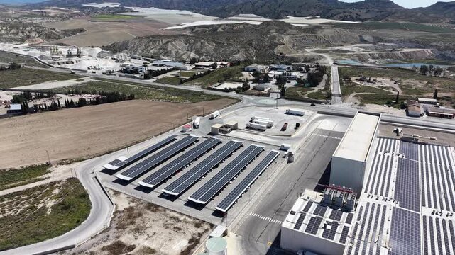 An orbiting drone shot of a modern logistics center featuring extensive solar energy installations on rooftops and parking lots in a dry valley.
