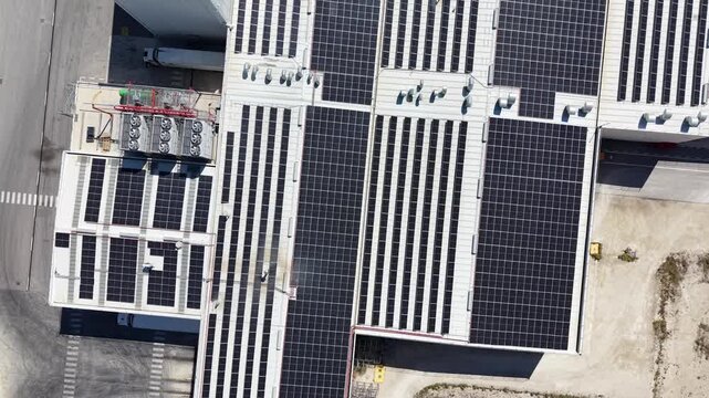 A top-down tracking drone shot moving across the roof of a large factory covered in solar panels, showcasing industrial-scale renewable energy.