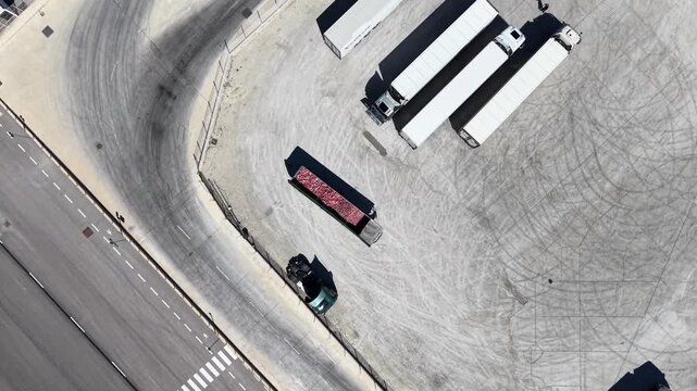 A top-down tracking drone shot following the movement of heavy trucks and trailers through a security checkpoint at an industrial facility in Spain.