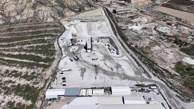 An orbiting drone shot around a gravel and asphalt business, showing the dusty yard, trucks, and mixing silos in the Murcia desert landscape.