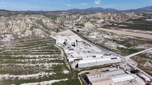An orbiting drone shot pulling back to reveal a cement factory nestled within the arid, desert-like hills and badlands of the Lorca region in Spain.