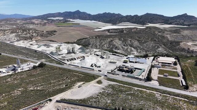 A tracking drone shot approaching a gravel and cement production facility, showcasing silos, piles of raw materials, and heavy machinery in the Murcia desert.