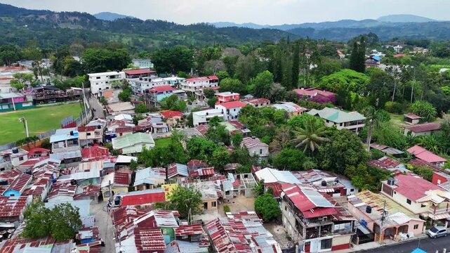 Aerial flyover of Jarabacoa, Dominican Republic, showing dense residential neighborhood with rusty metal rooftops, palm trees and nearby baseball field, surrounded by lush tropical hills and greenery.