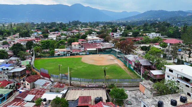 Aerial view of baseball field in Jarabacoa, Dominican Republic, surrounded by residential neighborhood and lush mountains. Local sports culture and community life in tropical setting. Low income.