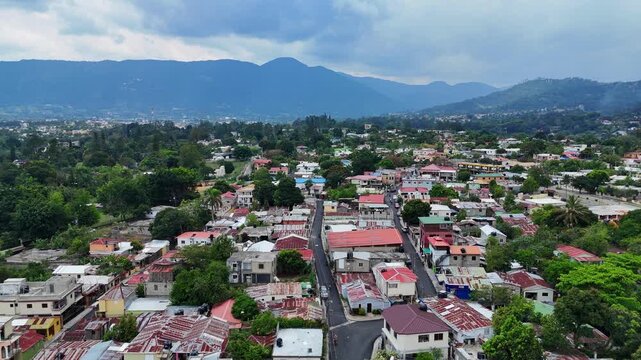 Aerial view of residential neighborhood in Jarabacoa, Dominican Republic, with modest homes, dense layout and lush green surroundings with mountains in background under cloudy daylight