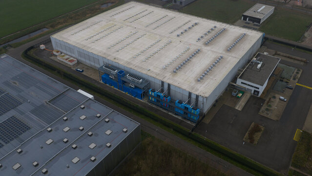 Roosendaal, Netherlands - 27 December 2025: Aerial view of a massive datacenter, its light roof contrasting with deep blue cooling units and dark green fields.