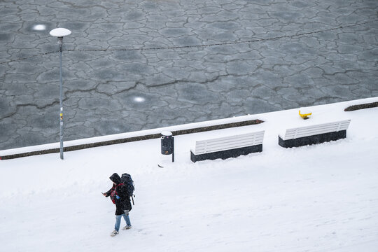 Tallinn, Estonia - 03 January 2025: View of a lone traveler trudging through crisp white snow alongside a frozen, fractured waterway, under the muted winter light.