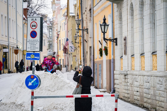 Tallinn, Estonia - 03 January 2025: View of a vibrant child playing on pristine snow, contrasting with the muted, historic street facades and festive lamps.