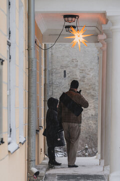 Tallinn, Estonia - 03 January 2025: View of two figures pausing under a portico, bathed in the warm glow of a star decoration, against the stark, snow-dusted path and ancient stone walls.
