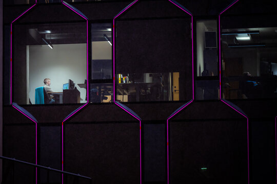 Tallinn, Estonia - 03 January 2025: View of a solitary figure bathed in warm light within a stark, dark modern building, its geometric windows outlined in vibrant magenta against the night.