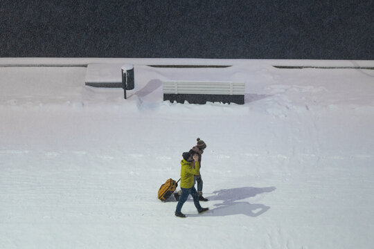 Tallinn, Estonia - 03 January 2025: View of two figures, one in a vibrant yellow jacket, trudge through a pristine snowscape, their shadows stark against the dark wall and snow-dusted bench.