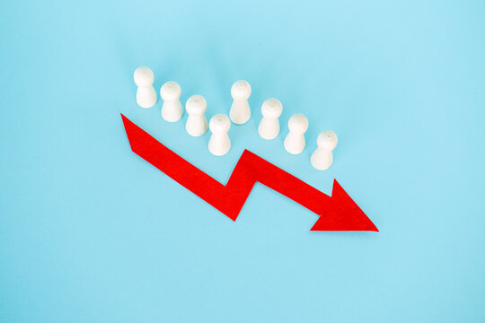 Line of wooden peg dolls stands behind red downward arrow on blue background. Population shrinking statistics, fertility rate drop, societal aging impact, Human capital depletion.