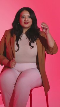 Vertical video: Starting, mid-adult woman adjusting hair while posing on stool in pink set, blazer