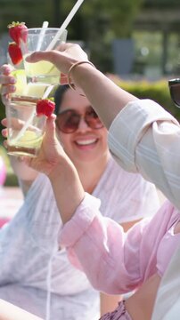 Lead woman lifting strawberry cocktail celebrating as friends clink and sip poolside for marketing