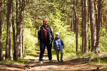 Fototapeta premium A father and his young son stand together on a dirt path in a sun-drenched pine forest, looking ahead during a family hike or nature walk.