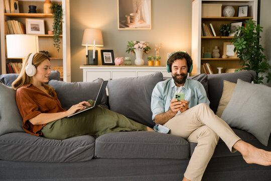 Two people relaxing on a sofa using laptop and mobile phone with headphones
