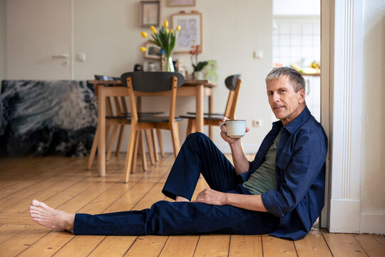 Senior man with coffee cup sitting on hardwood floor at home