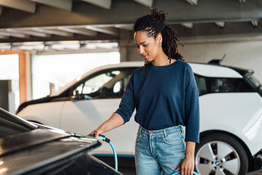 Smiling young businesswoman charging electric car at parking garage
