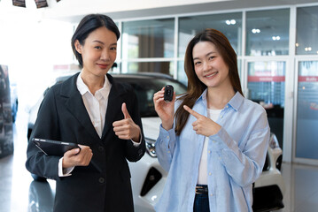 Obraz premium Successful car purchase. Happy Asian woman receiving new car keys from a professional dealer in a showroom. Automotive sales and excellent service concept.