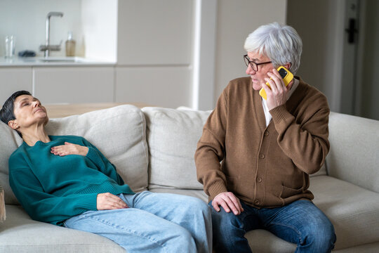 Worried concerned senior husband urgently calling to ambulance while elderly wife lying on sofa having heart attack. Aged woman collapsed on couch with chest pain, man checks on wife and calls doctor.