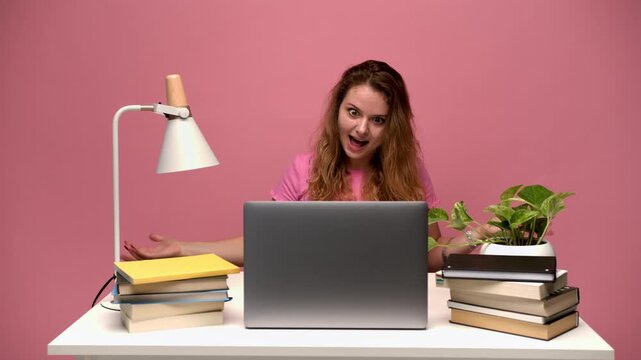 Angry student shouting and gesturing at her laptop computer while studying online at her desk with a pink background