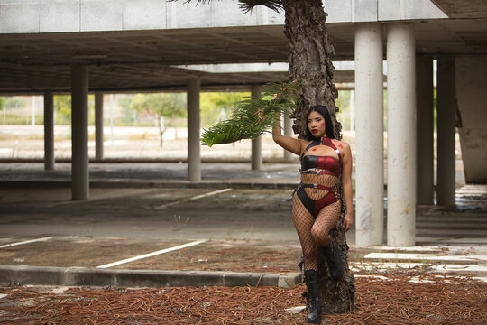 Young Latina woman, brunette, red eyes, wearing a leather suit and boots, fishnet stockings, posing leaning against a tree trunk in a post-apocalyptic city, as if she had become a zombie.
