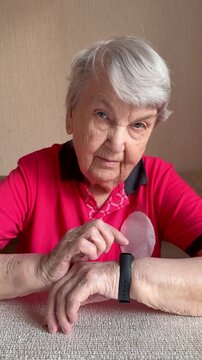 Elderly woman checking watch on wrist while seated at table tapping screen of fitness band, focused expression, red polo shirt
