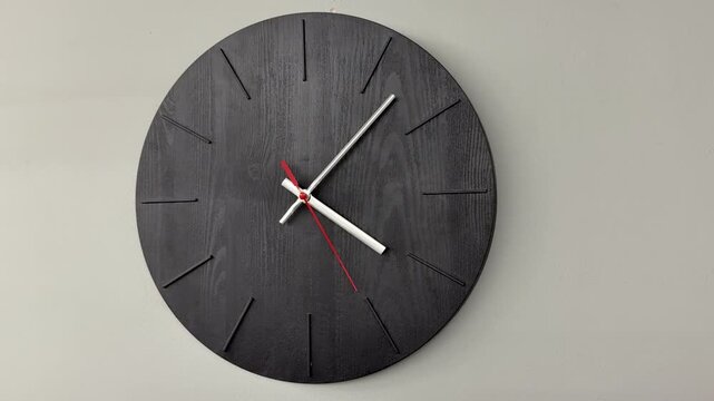 Black wooden wall clock on grey wall, minimalist timepiece with textured round face, white hour and minute hands, red second hand