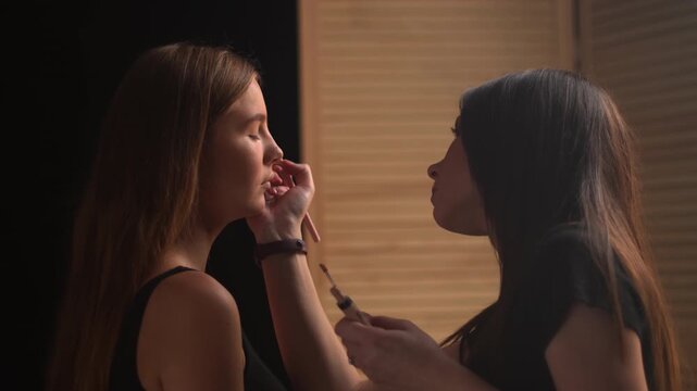 Professional makeup artist applying foundation with a brush to a female model's nose in a beauty salon, meticulously preparing her skin for a perfect complexion