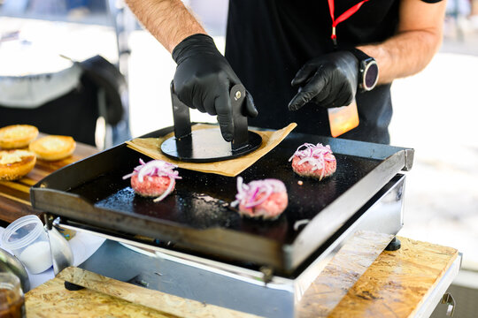 Chef preparing smash burgers on a flat top grill, adding toppings to freshly cooked beef patties with melted cheese, capturing street food cooking process and professional burger preparation in a