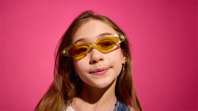 Child girl in yellow sunglasses looking at camera on pink background with clean studio light and minimal composition. Concept of eyewear promo, kids branding, advertising content.