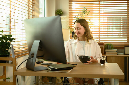 Young Caucasian woman enjoying breakfast cereal while working at her desk in a cozy home office