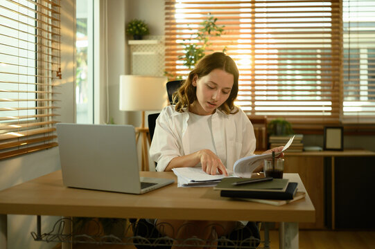 Young Caucasian woman working at home, reviewing documents while using a laptop in a bright and comfortable office
