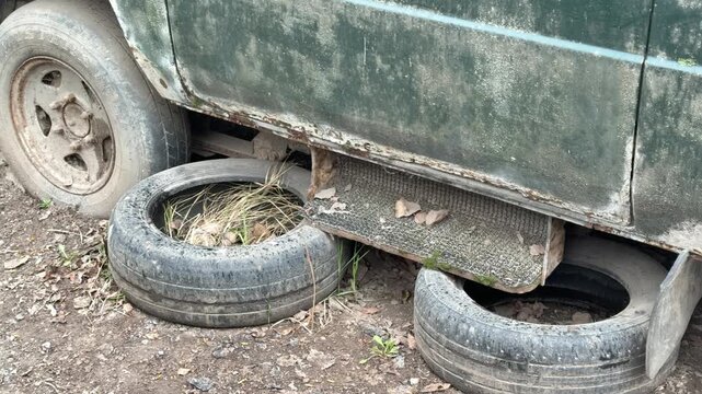 Abandoned dark green minibus punctured tire, rusted undercarriage and chipped paint, spare tires stacked beneath body, moss.
