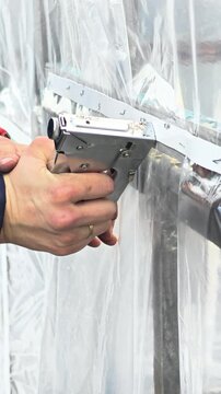 Hands using a staple gun to secure plastic sheeting. The scene focuses on construction or renovation work, emphasizing tools and materials.