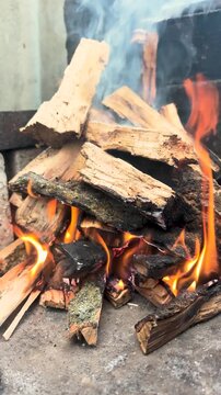 A close-up of a burning firewood stack with flames and smoke rising. The wood is rough and varied in size, creating a warm, inviting atmosphere.