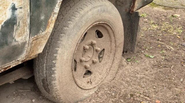 Abandoned dark green minibus with punctured tire, close-up of muddy wheel and spare rim tucked under chassis, heavy rust patches