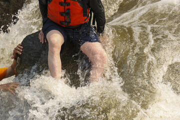 Fototapeta premium Adventurous person wearing bright orange life jacket sits rock amidst fast flowing whitewater river rapids expressing intense thrill and determination during exciting outdoor water sport teamwork