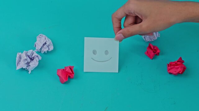 Hand placing smiling sticky note over sad face cutout surrounded by crumpled paper
