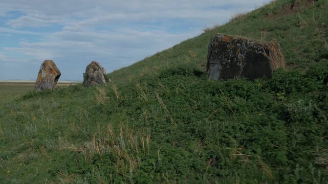 Handheld shot viewing ancient megalithic standing stones embedded in the green, grassy slope of a kurgan burial mound. The historic archaeological landscape is set against a cloudy summer sky.