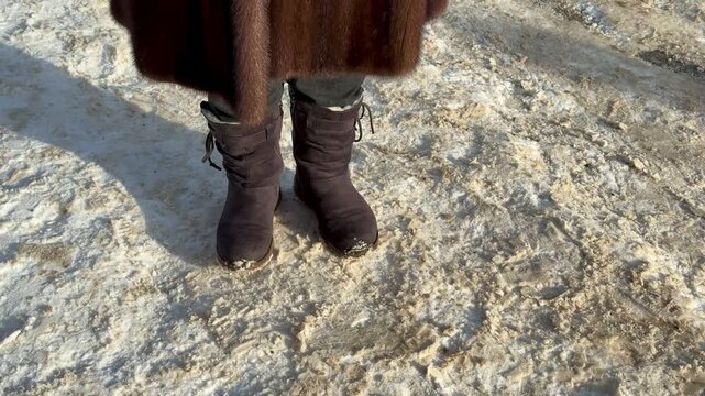 Mink coat suede boots in snow. Woman shifting weight, stomping feet to warm, visible breath in frosty air, sunlight highlighting