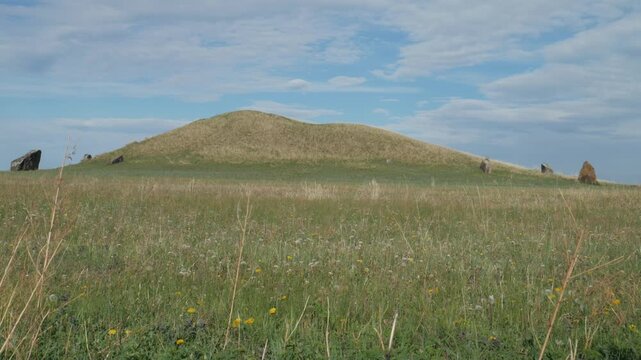 Handheld static wide shot of an ancient kurgan burial mound surrounded by megalithic standing stones. The historic grassy hill sits in a green summer meadow under a cloudy blue sky.