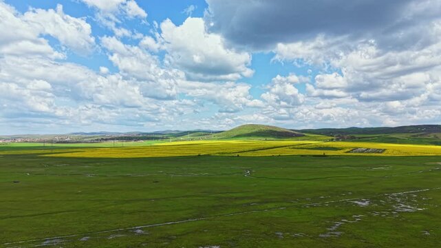 Green fields and yellow flowers cover the land under a bright blue sky with fluffy clouds. The scene shows hills in the background and a vast open area.