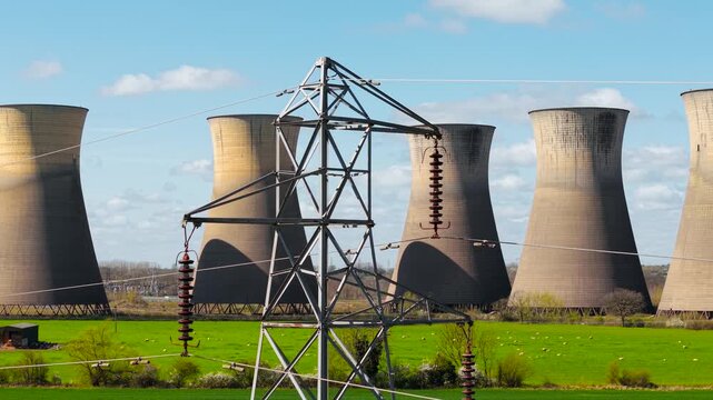 Steel power line tower with ceramic insulators near nuclear power plant cooling towers and green pasture. High voltage electricity transmission with rural landscape and atomic energy facility. Grid